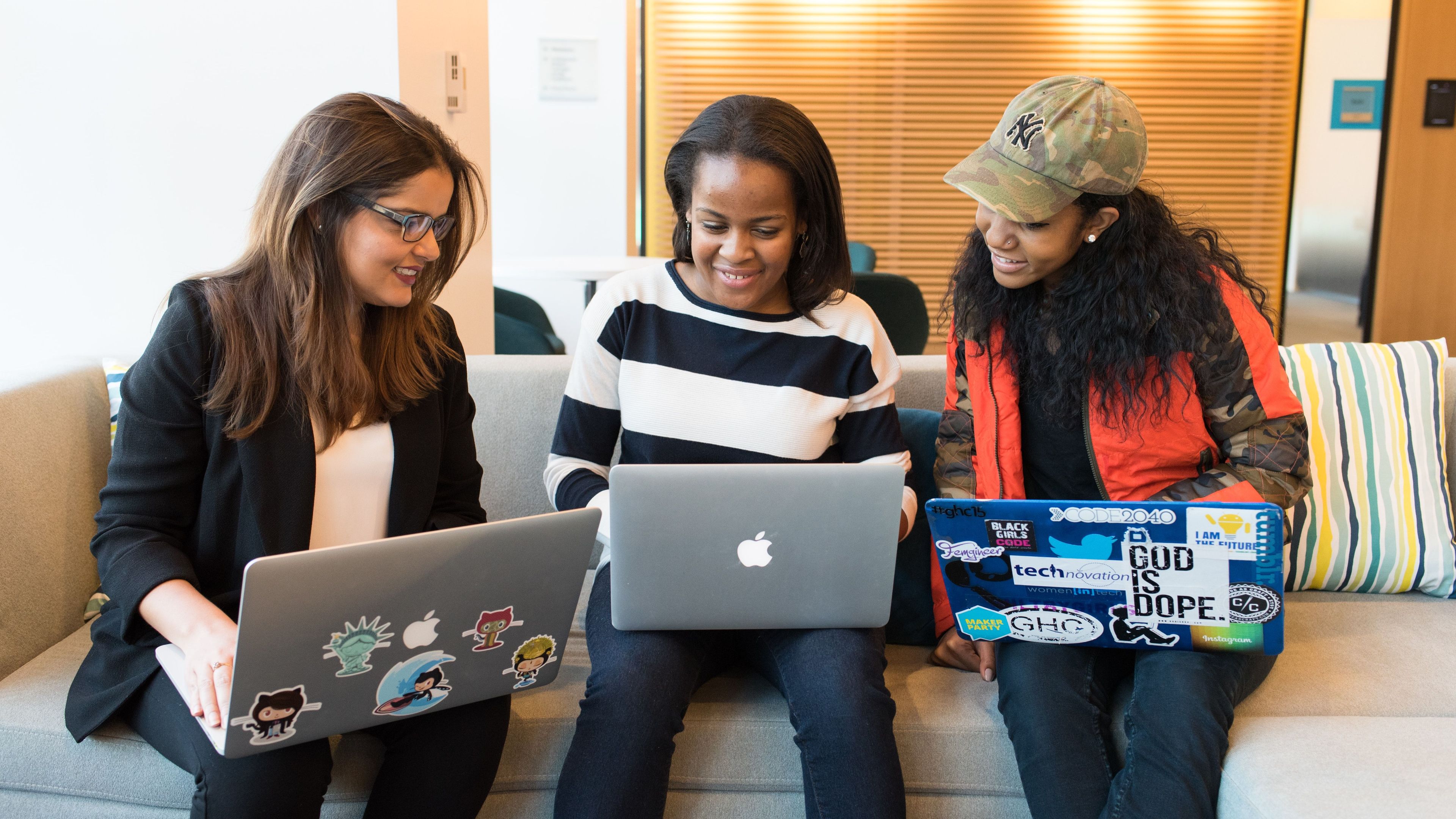 Three women sitting on a couch, using laptops decorated with stickers, and smiling.