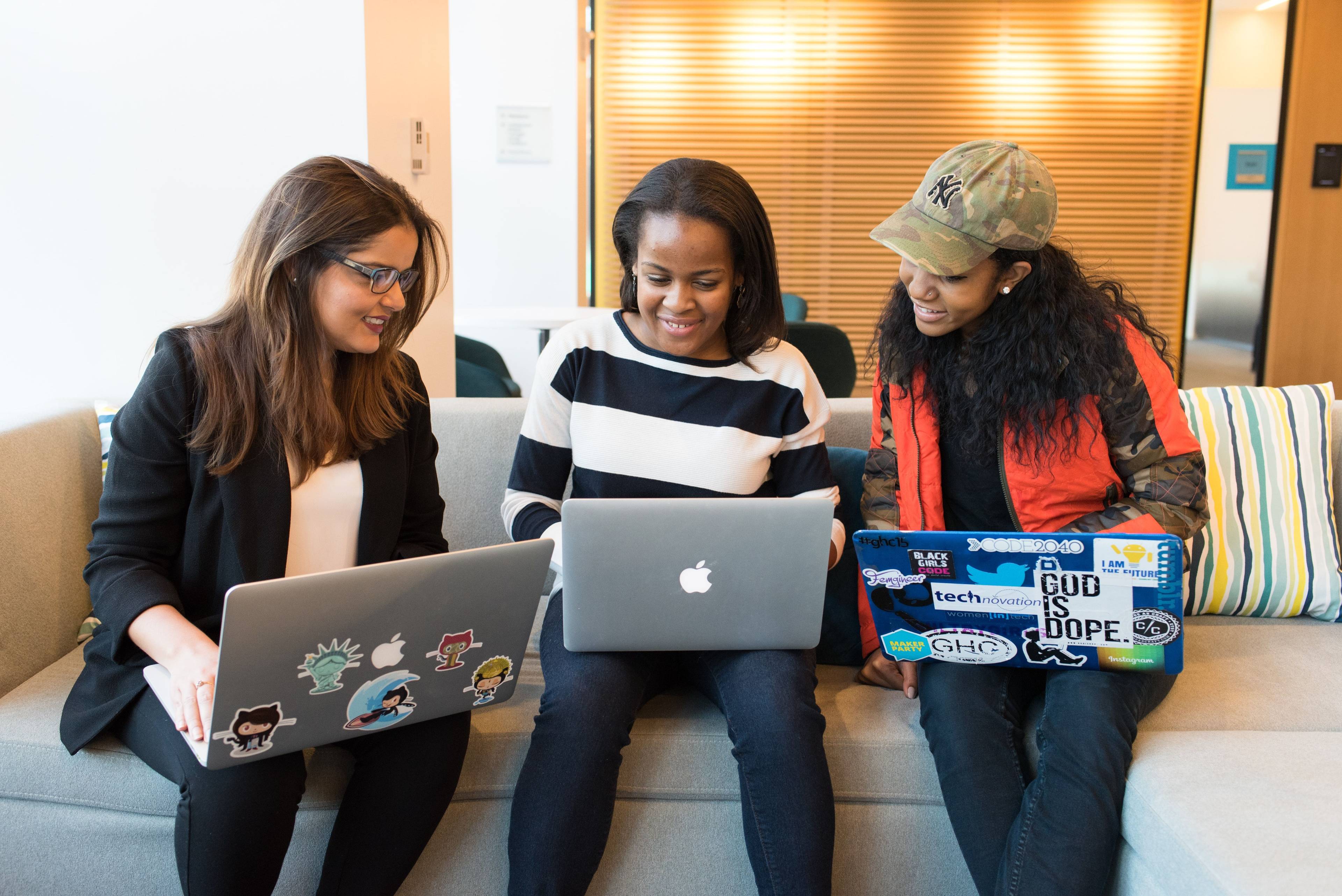 Three women sitting on a couch, using laptops decorated with stickers, and smiling.