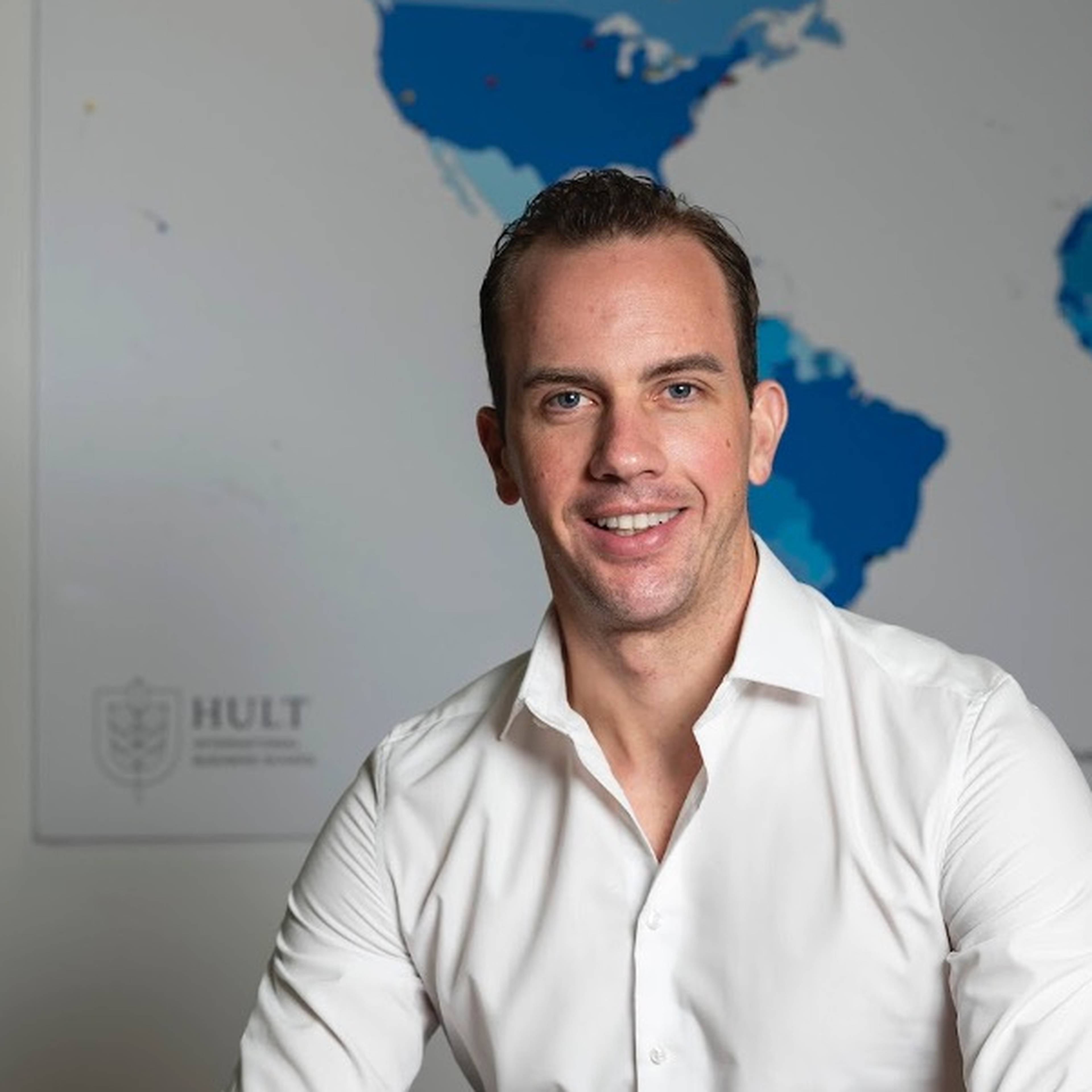Man in white shirt sits and smiles in front of world map.