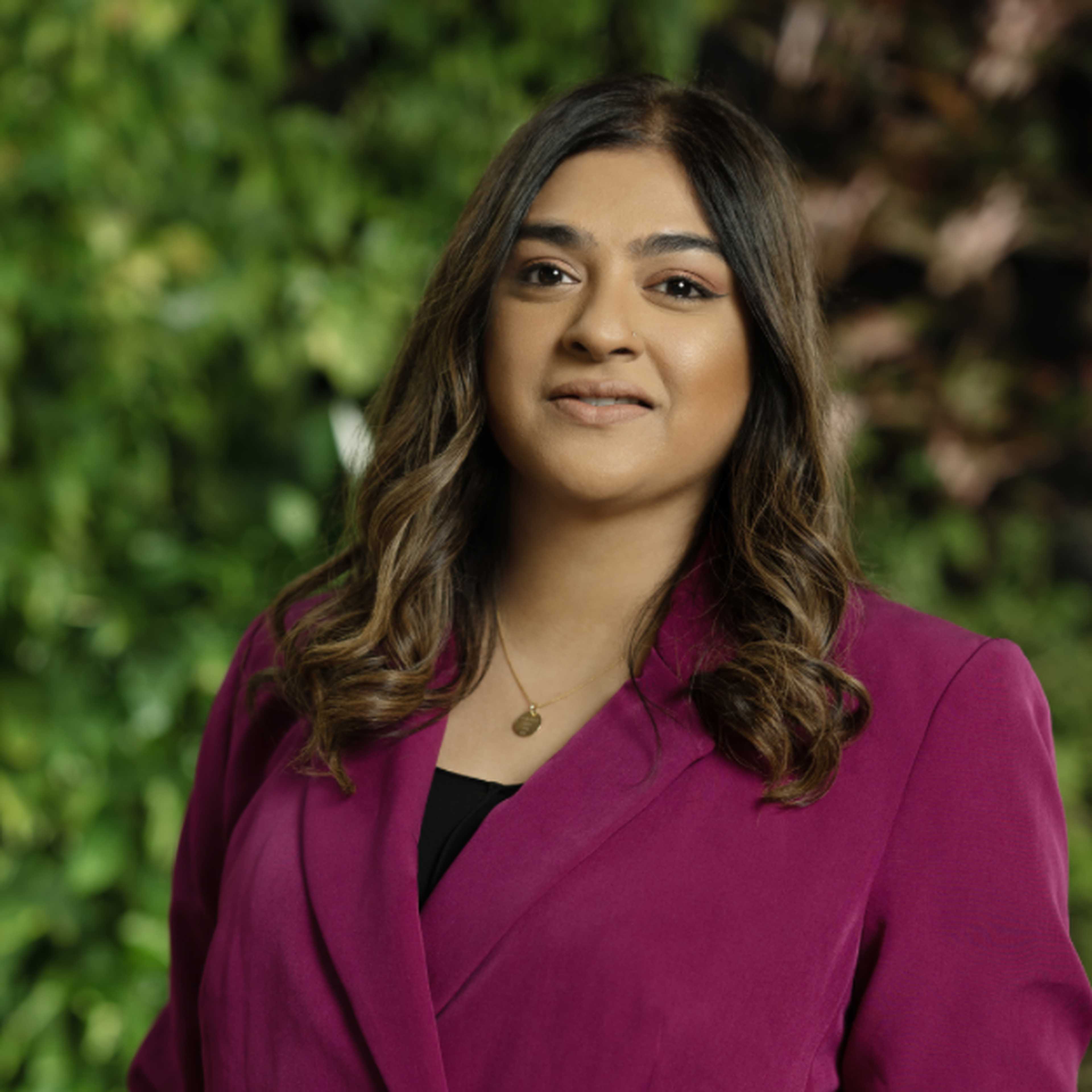 A woman in a magenta blazer stands confidently with a blurred green foliage background.