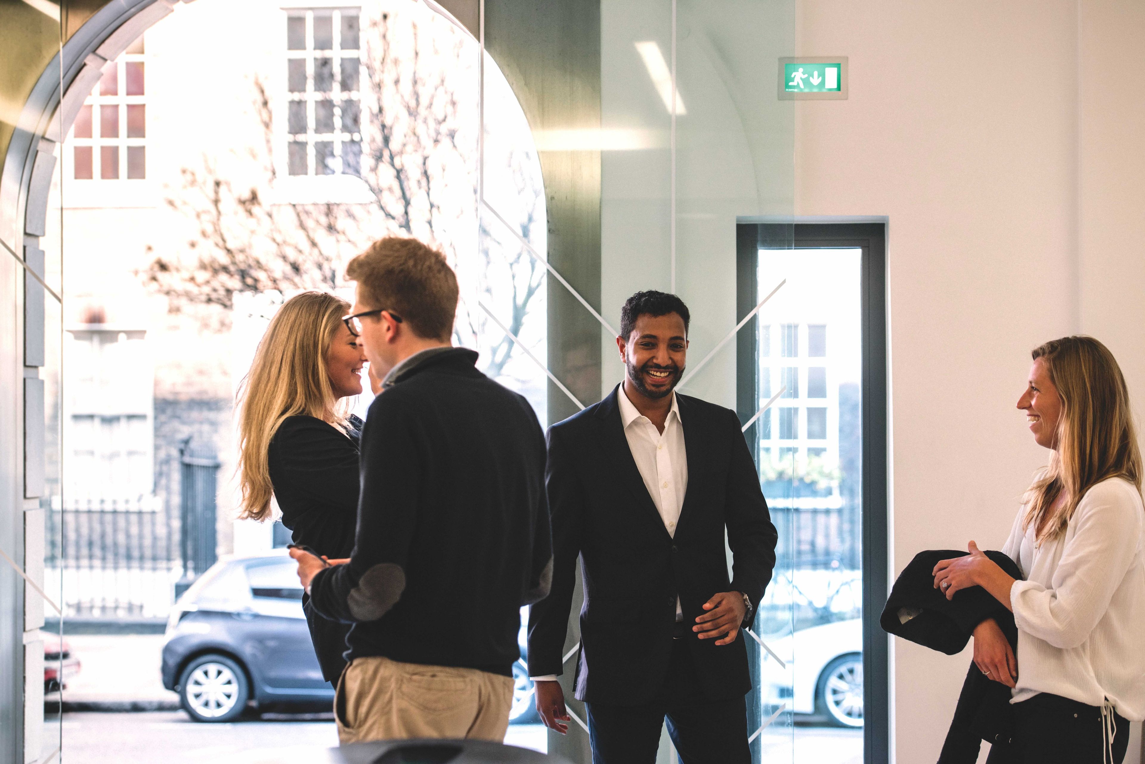 Four people in business attire interact and smile inside a modern office with large windows and an exit sign visible.