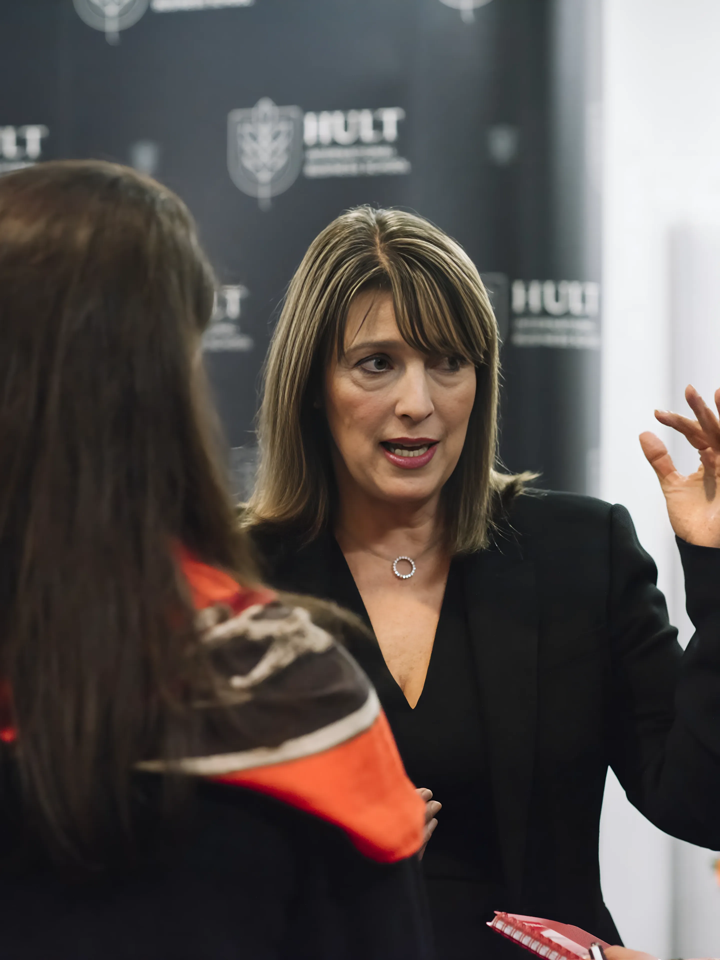 A woman in a black blazer gestures while speaking to a small group indoors.