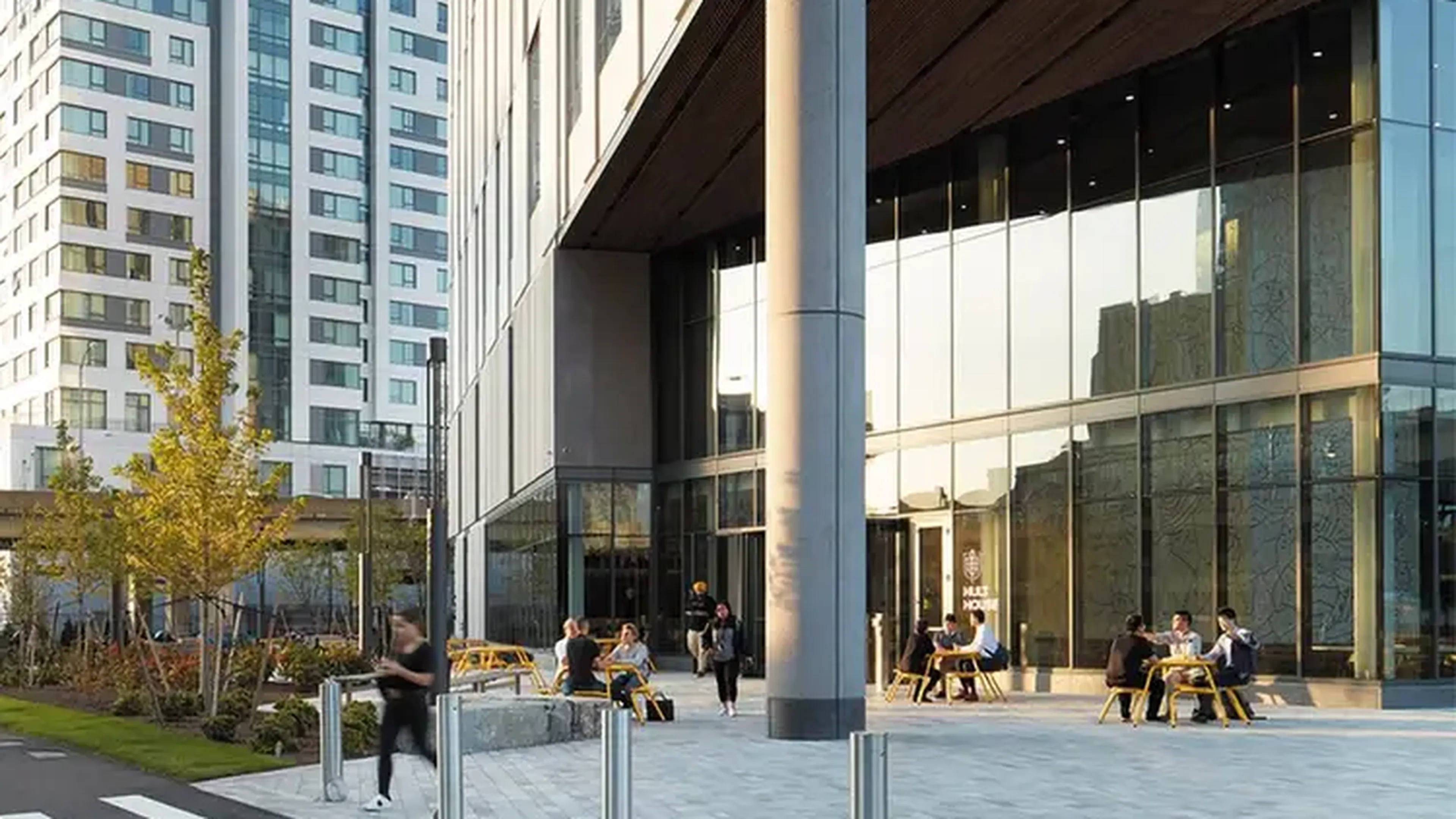 People sitting at outdoor tables in front of a modern glass building with urban surroundings.