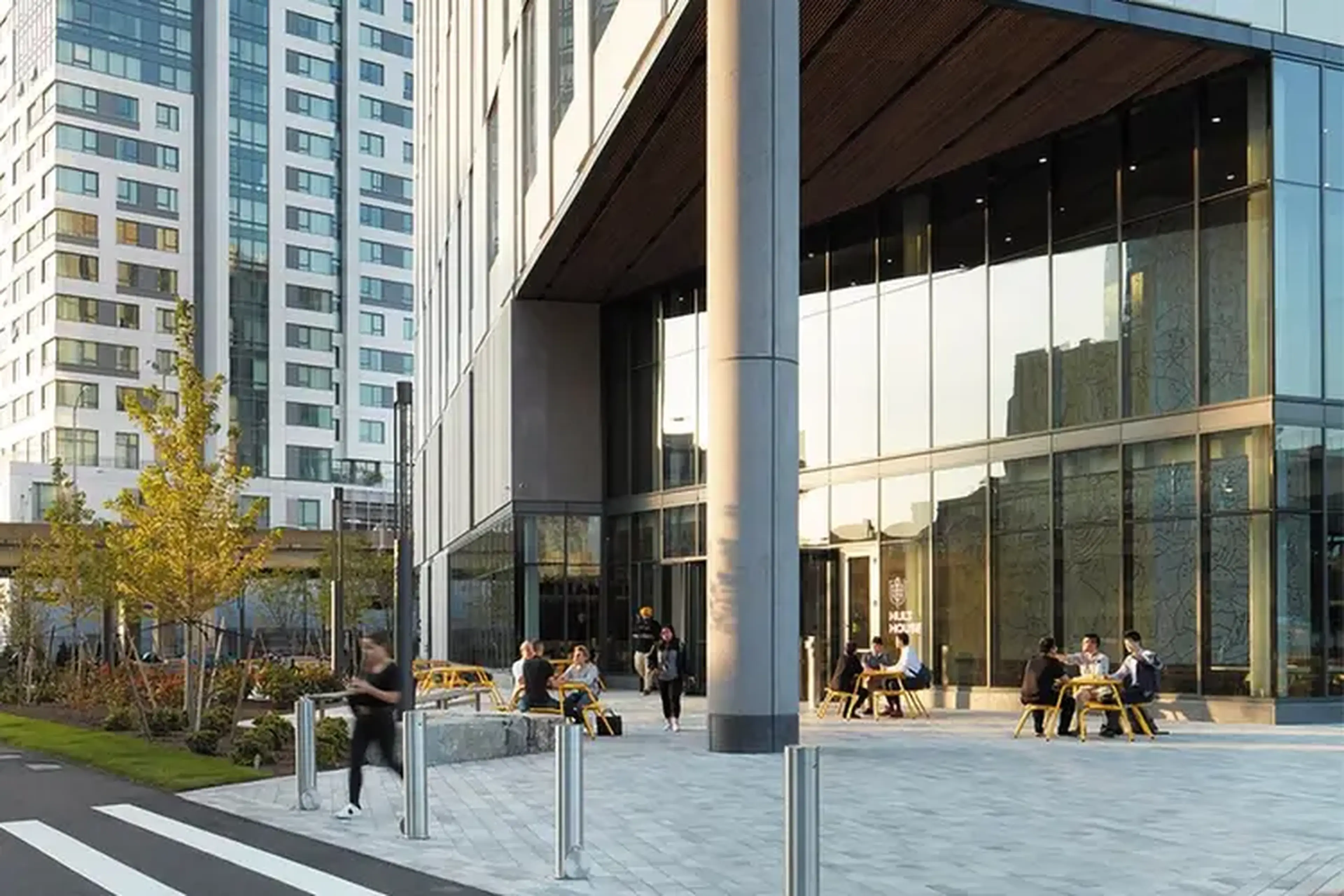 People sitting at outdoor tables in front of a modern glass building with urban surroundings.