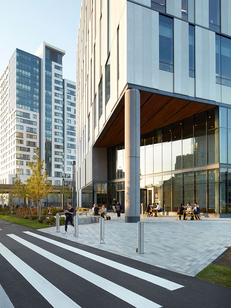 Modern glass building with people sitting outside, adjacent to a crosswalk, near tall residential towers and landscaped greenery.