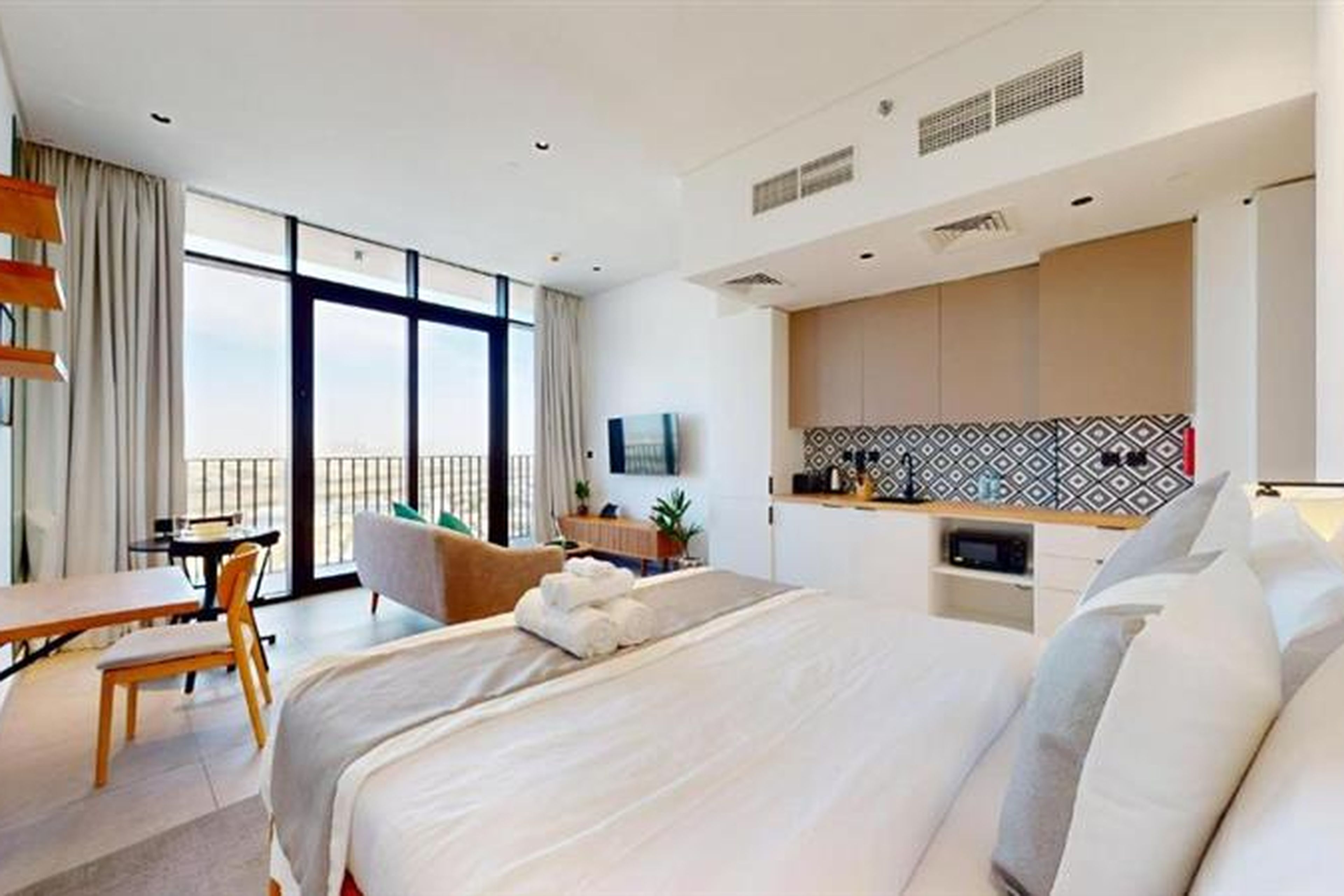 Modern studio apartment with a bed, kitchenette, and sitting area. Large windows and a balcony offer a bright view. Neutral tones and minimalist decor.
