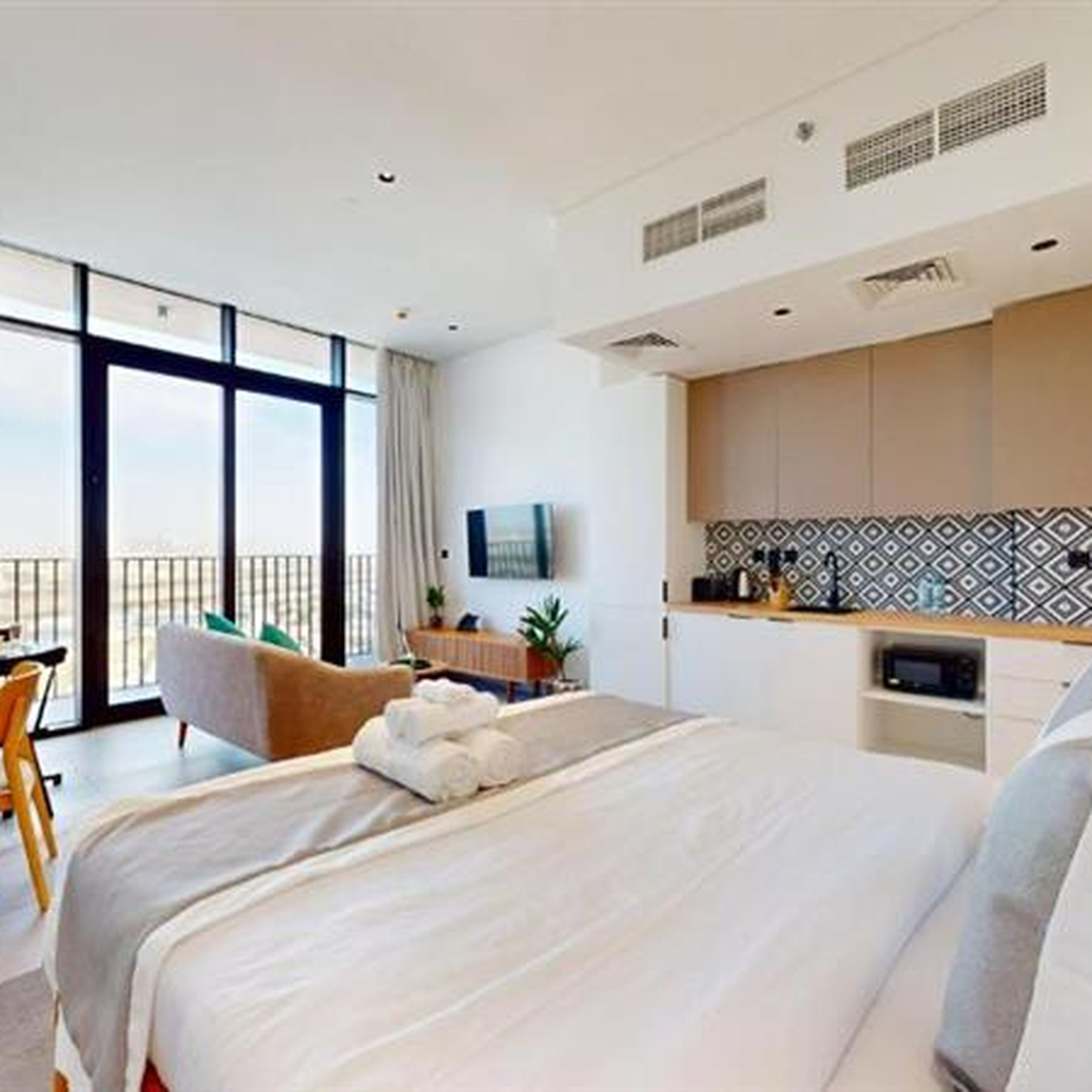 Modern studio apartment with a bed, kitchenette, and sitting area. Large windows and a balcony offer a bright view. Neutral tones and minimalist decor.