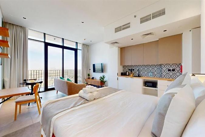 Modern studio apartment with a bed, kitchenette, and sitting area. Large windows and a balcony offer a bright view. Neutral tones and minimalist decor.