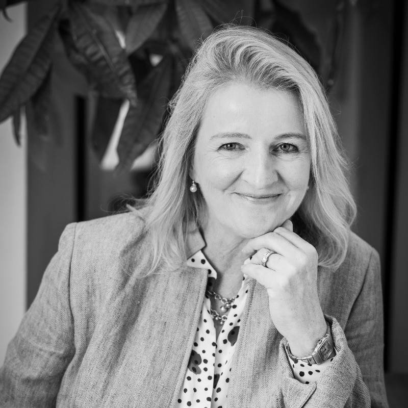 Smiling woman with blonde hair in a blazer, seated and resting chin on hand, indoors.