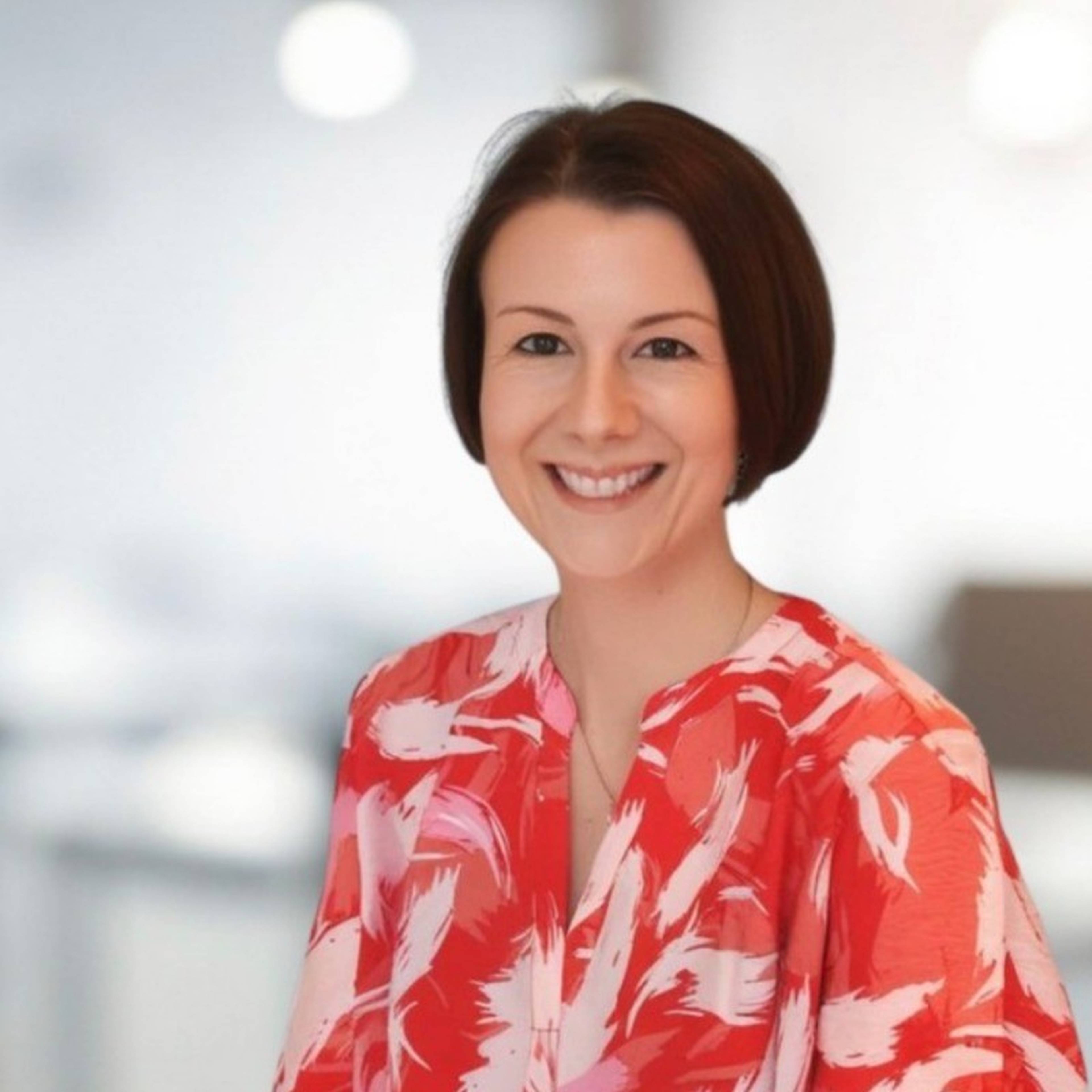 Smiling woman in a red and white patterned blouse, with short brown hair, in a softly blurred indoor setting.