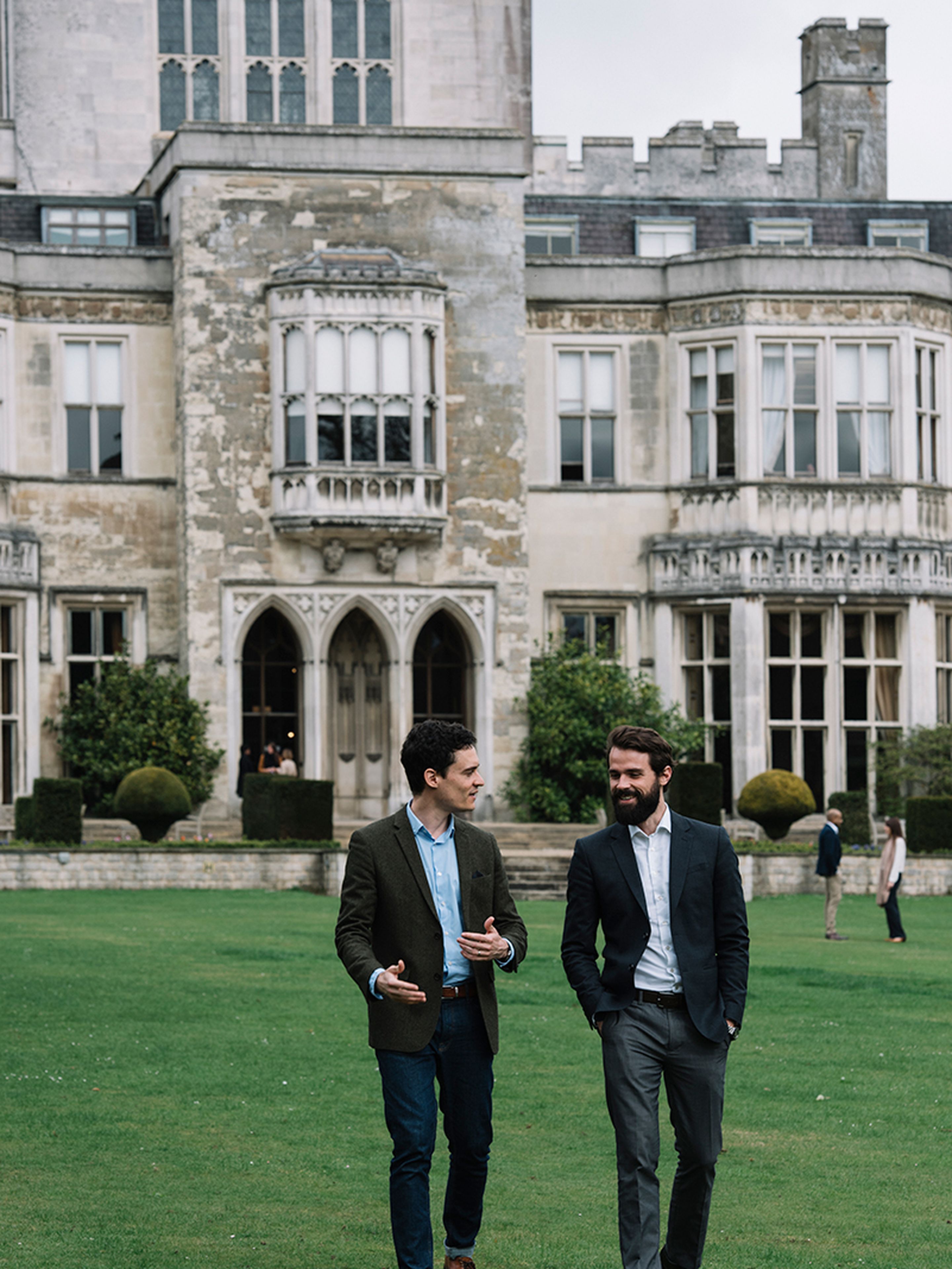 Two men in suits walking and talking on a lawn in front of a historic stone building with large windows and arched doorways.