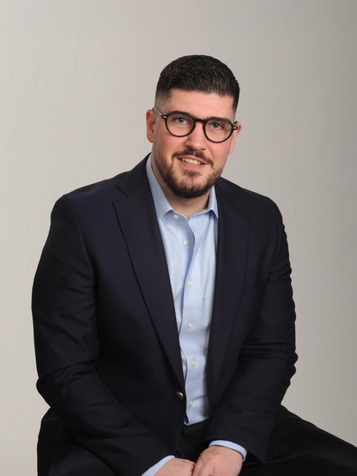 Man in a dark suit and glasses sitting against a neutral background, looking at the camera with a slight smile.
