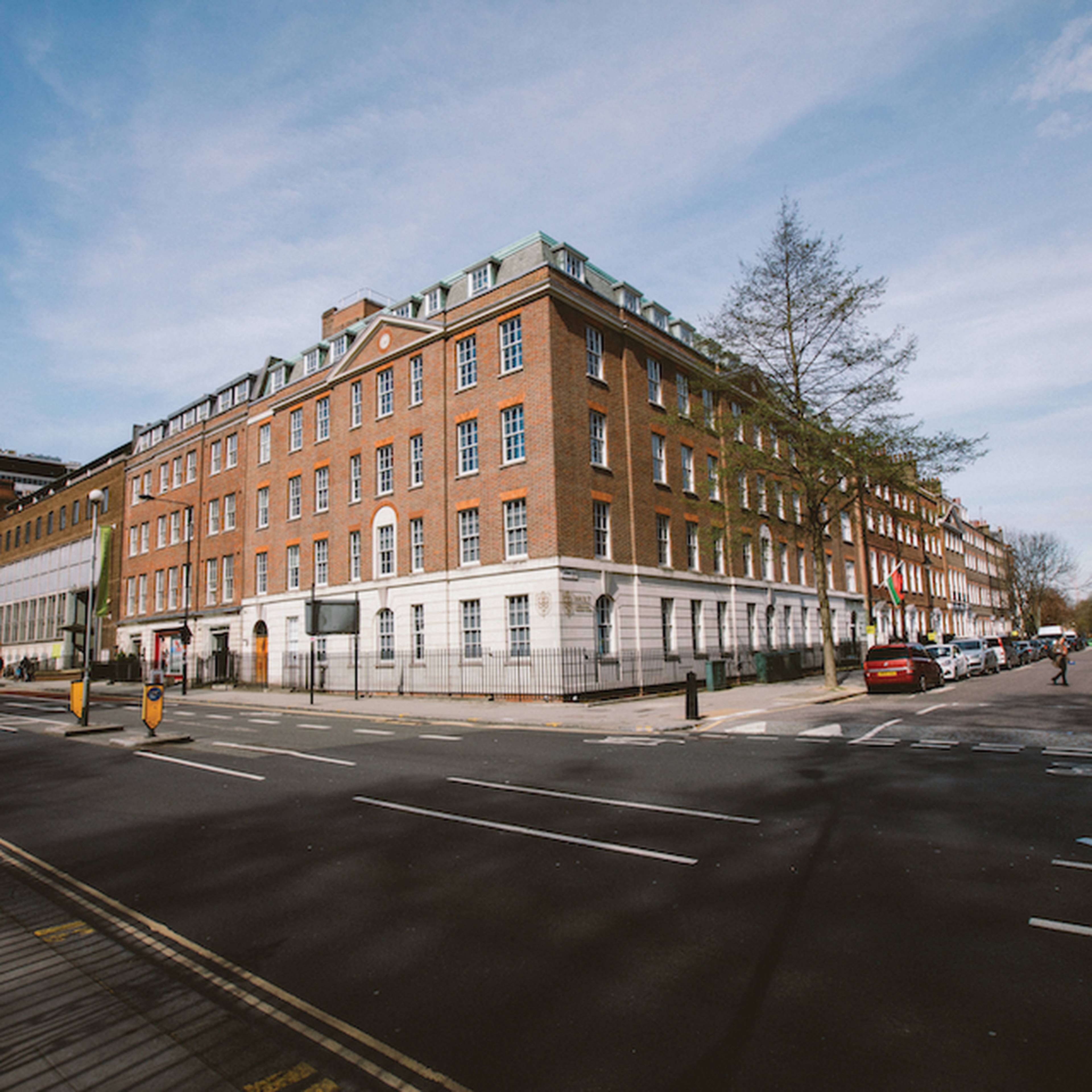 A red-brick building on a sunny street corner with parked cars and pedestrians.
