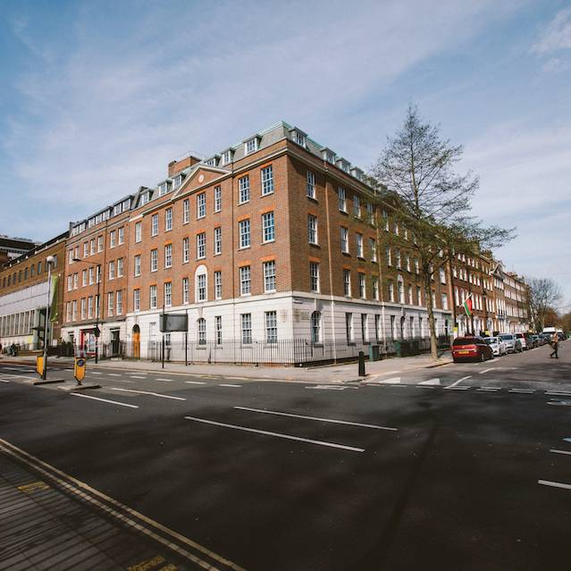 A red-brick building on a sunny street corner with parked cars and pedestrians.
