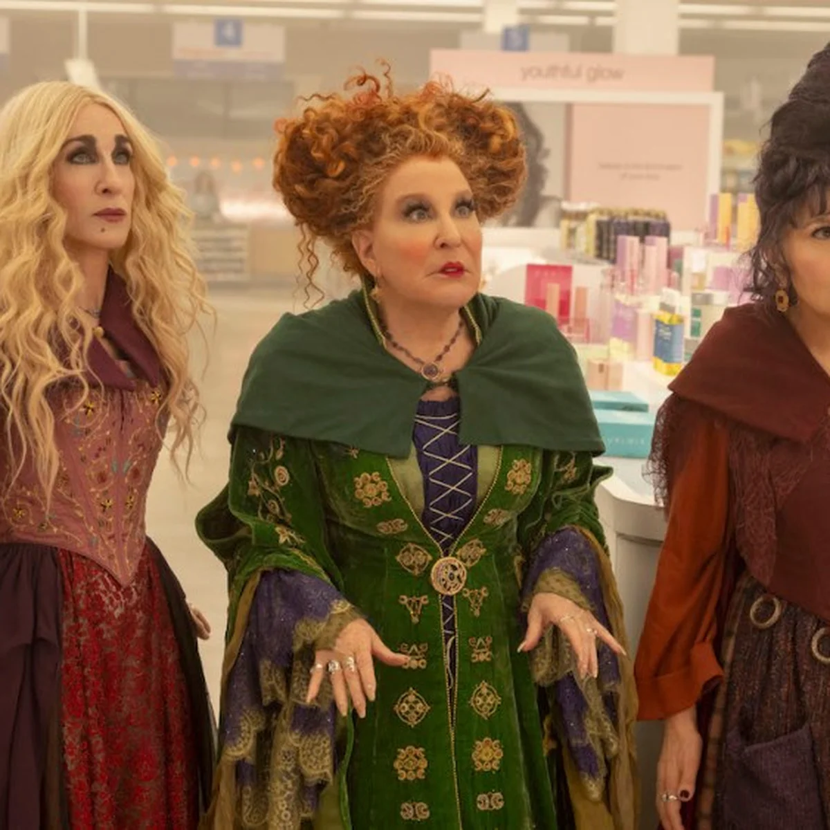 Three women in elaborate costumes inside a store, with shelves of products in the background. They appear to be surprised or curious.