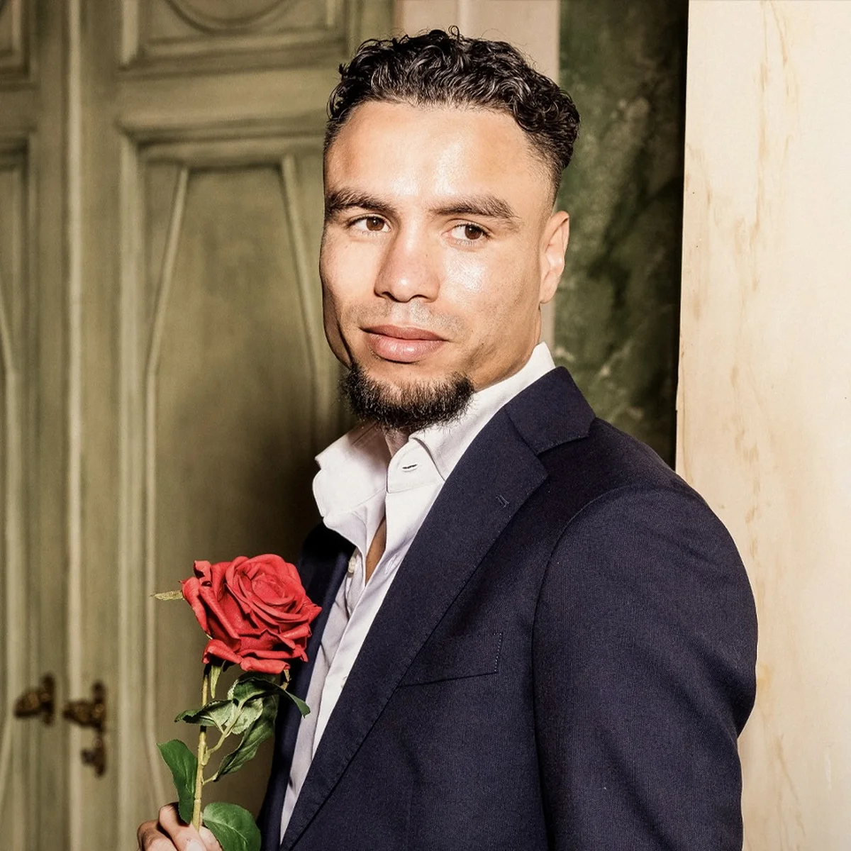 Man in a suit holding a red rose, standing against a textured wall with a slight smile.