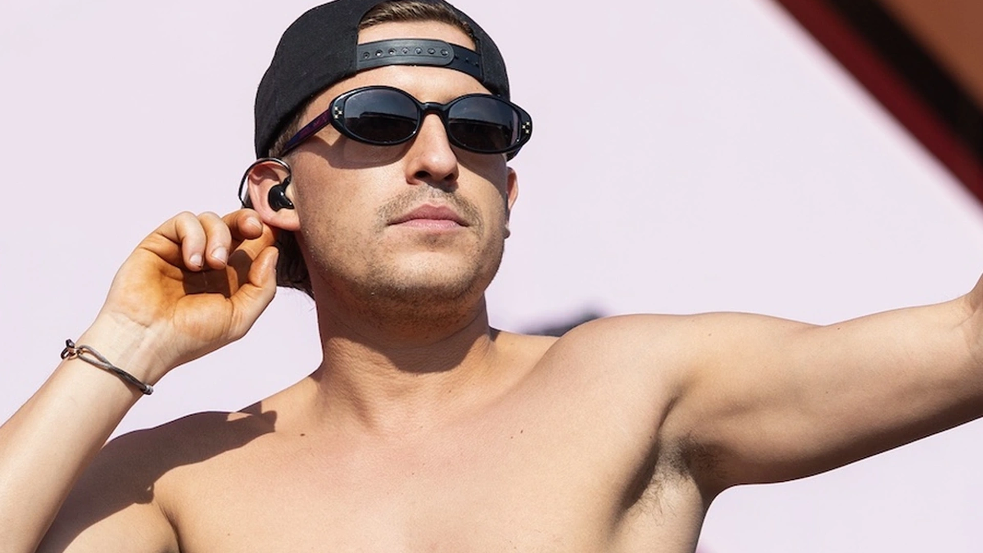 Shirtless man with sunglasses and backward cap, holding an in-ear monitor on stage, with tattoos on his chest.