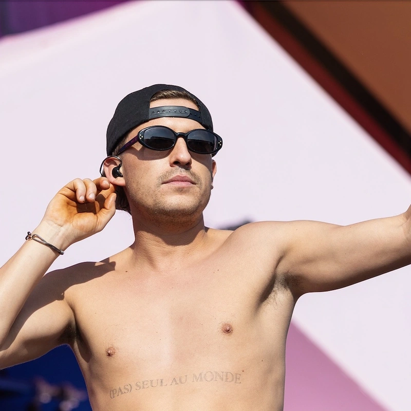 Shirtless man with sunglasses and backward cap, holding an in-ear monitor on stage, with tattoos on his chest.