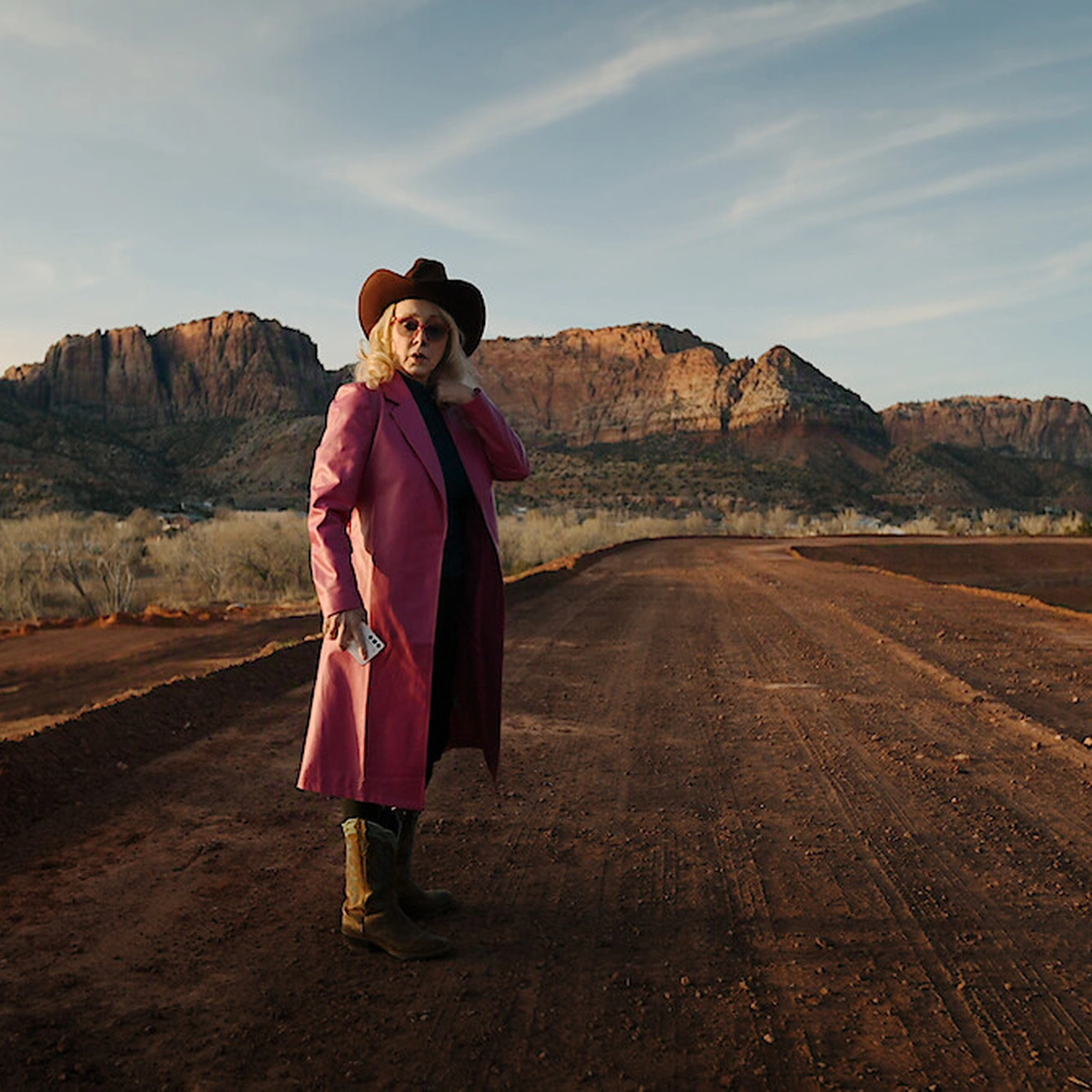 Person in a pink coat and cowboy hat stands on a dirt road with red rock cliffs in the background under a partly cloudy sky.