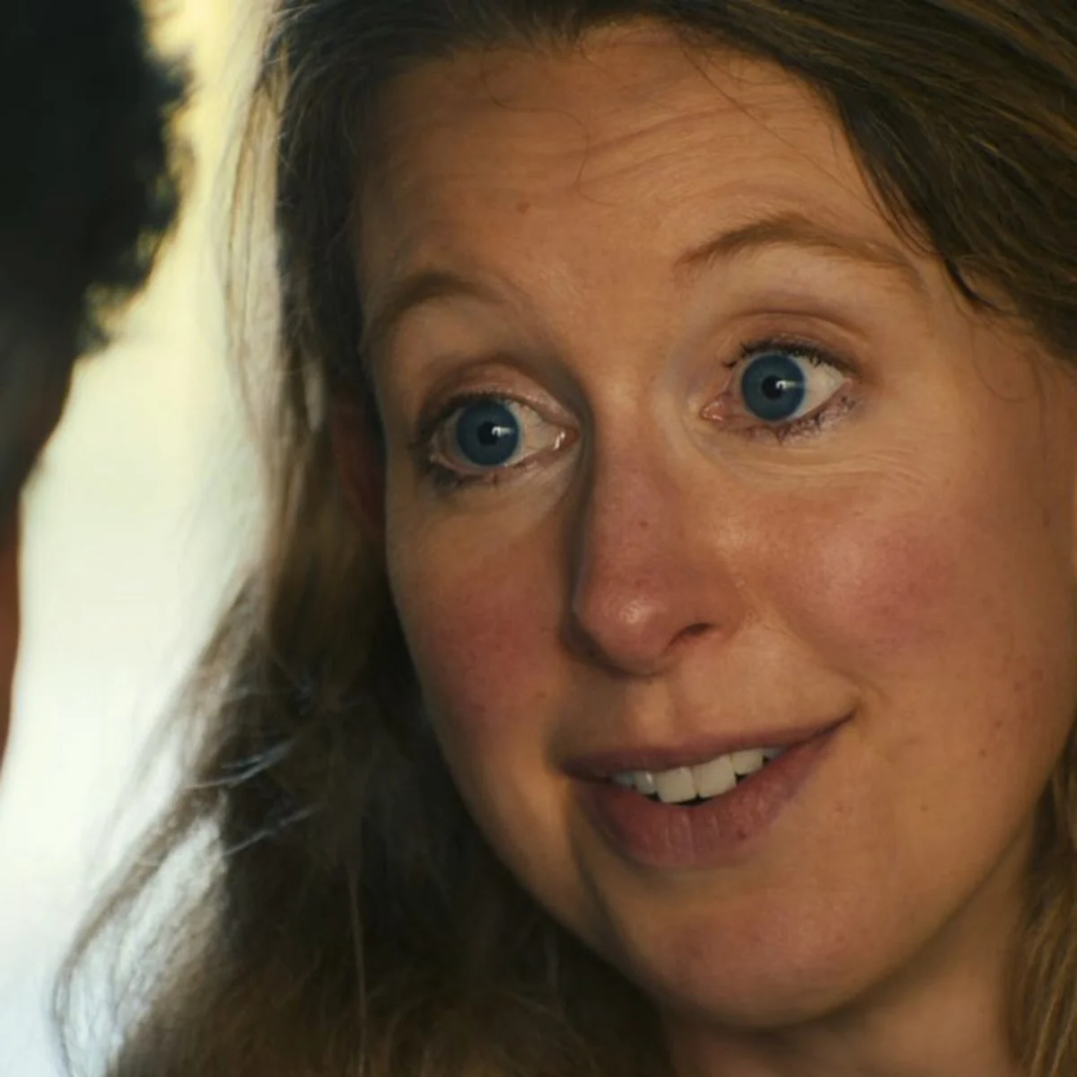 A woman with blue eyes and long hair is smiling and talking to a man with gray hair, who is turned away.