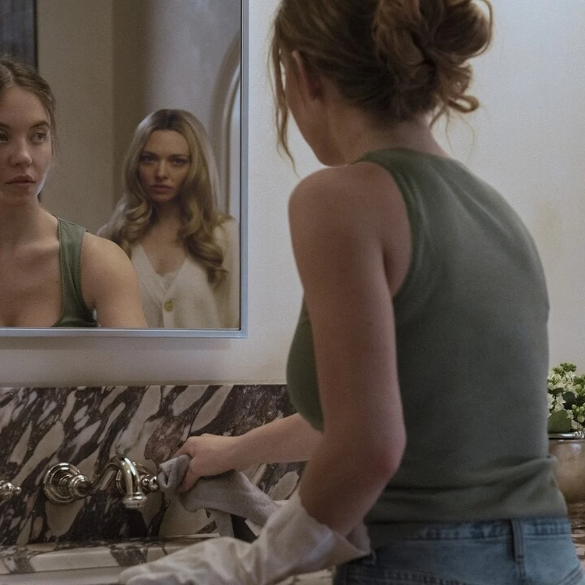 Woman cleaning a marble counter glances at her reflection in a mirror, where another woman is seen behind her.
