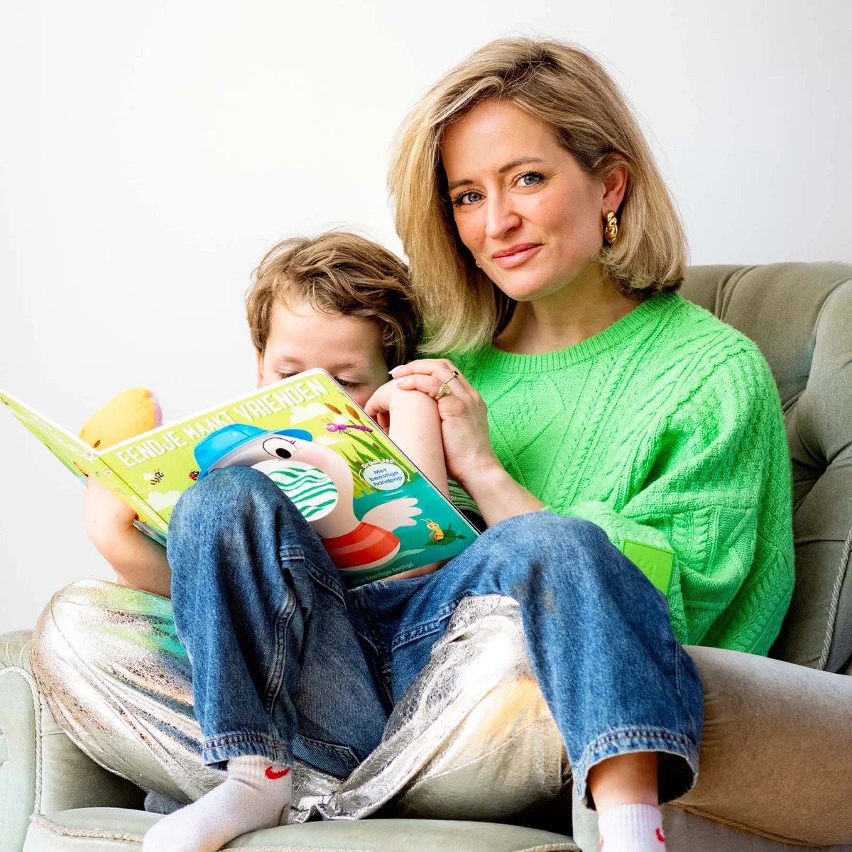 Woman in a green sweater sitting on a chair, holding a child reading a colorful book together.