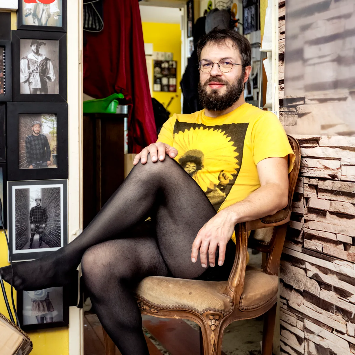 A bearded person in glasses, wearing a yellow shirt and black stockings, sits cross-legged on a chair in a room filled with framed photos.