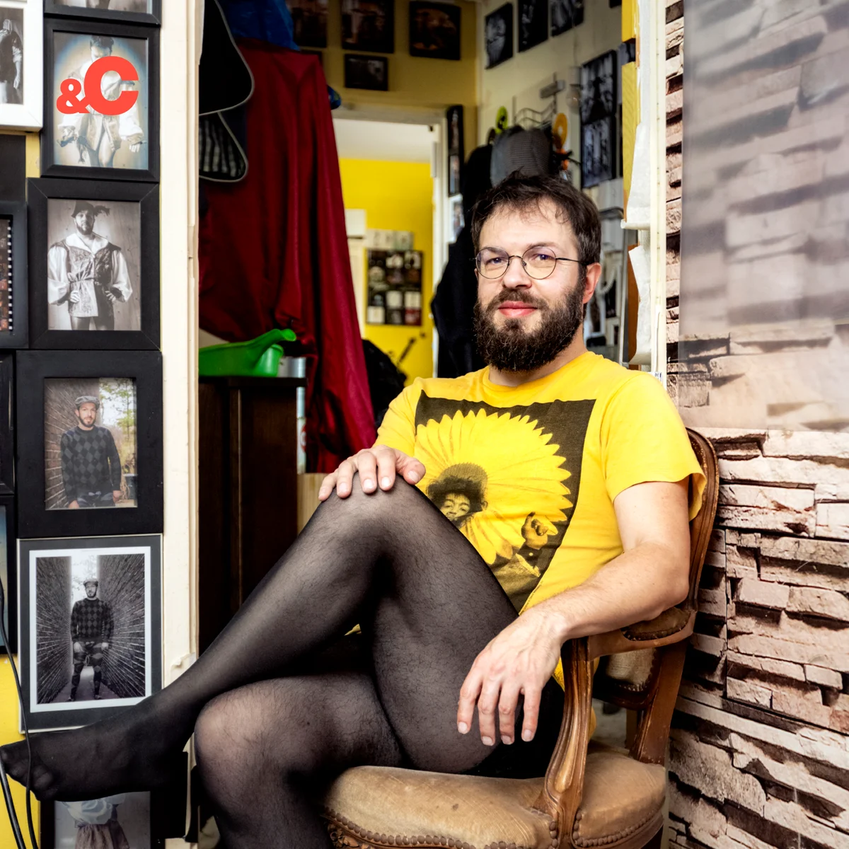 A bearded person in glasses, wearing a yellow shirt and black stockings, sits cross-legged on a chair in a room filled with framed photos.