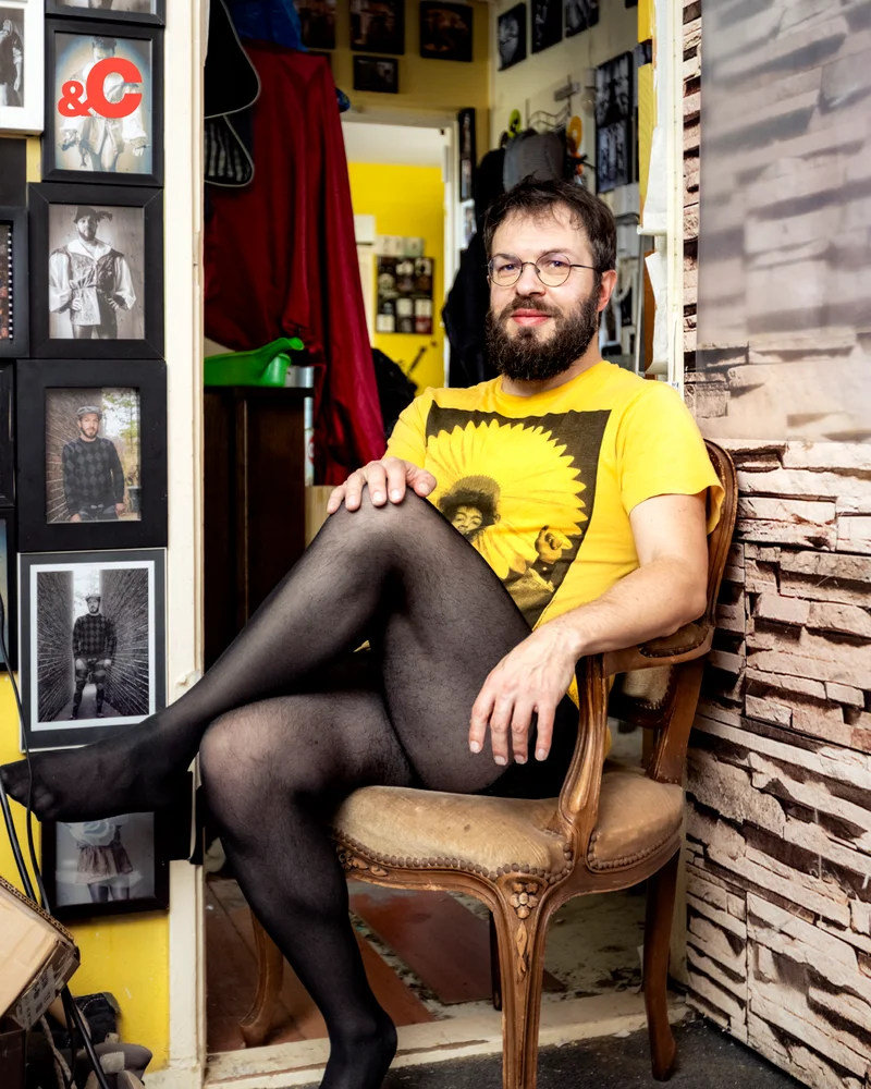 A bearded person in glasses, wearing a yellow shirt and black stockings, sits cross-legged on a chair in a room filled with framed photos.