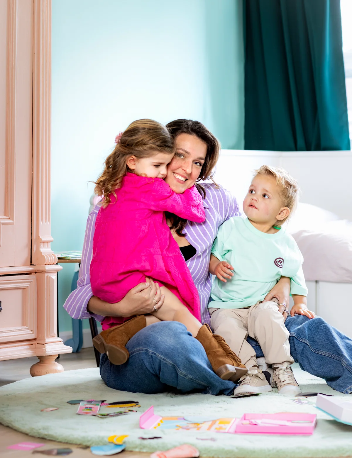 Woman hugging two children, one in pink, the other in mint, sitting on the floor in a bright room with toys scattered around.