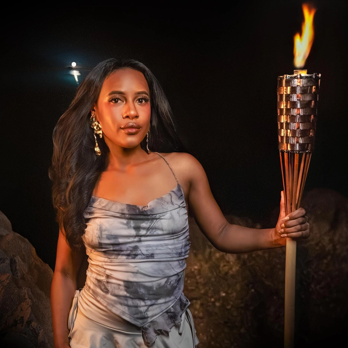 A woman holds a flaming torch at night, standing by rocks under the moonlight.