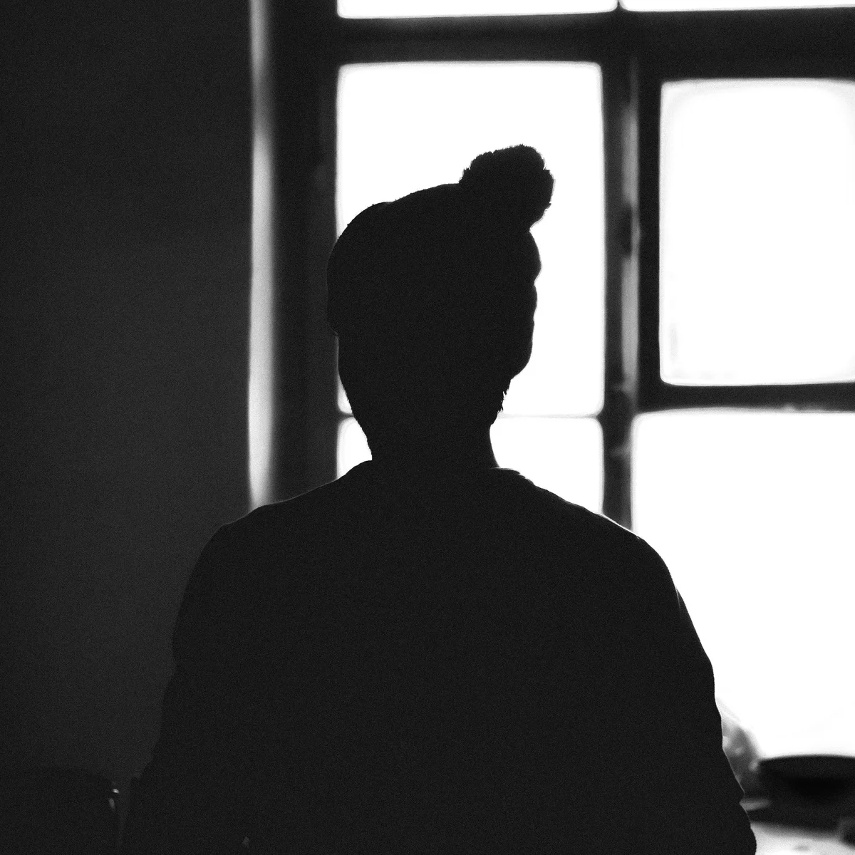 Silhouette of a person wearing a beanie, standing in front of a brightly lit window in a dimly lit room.