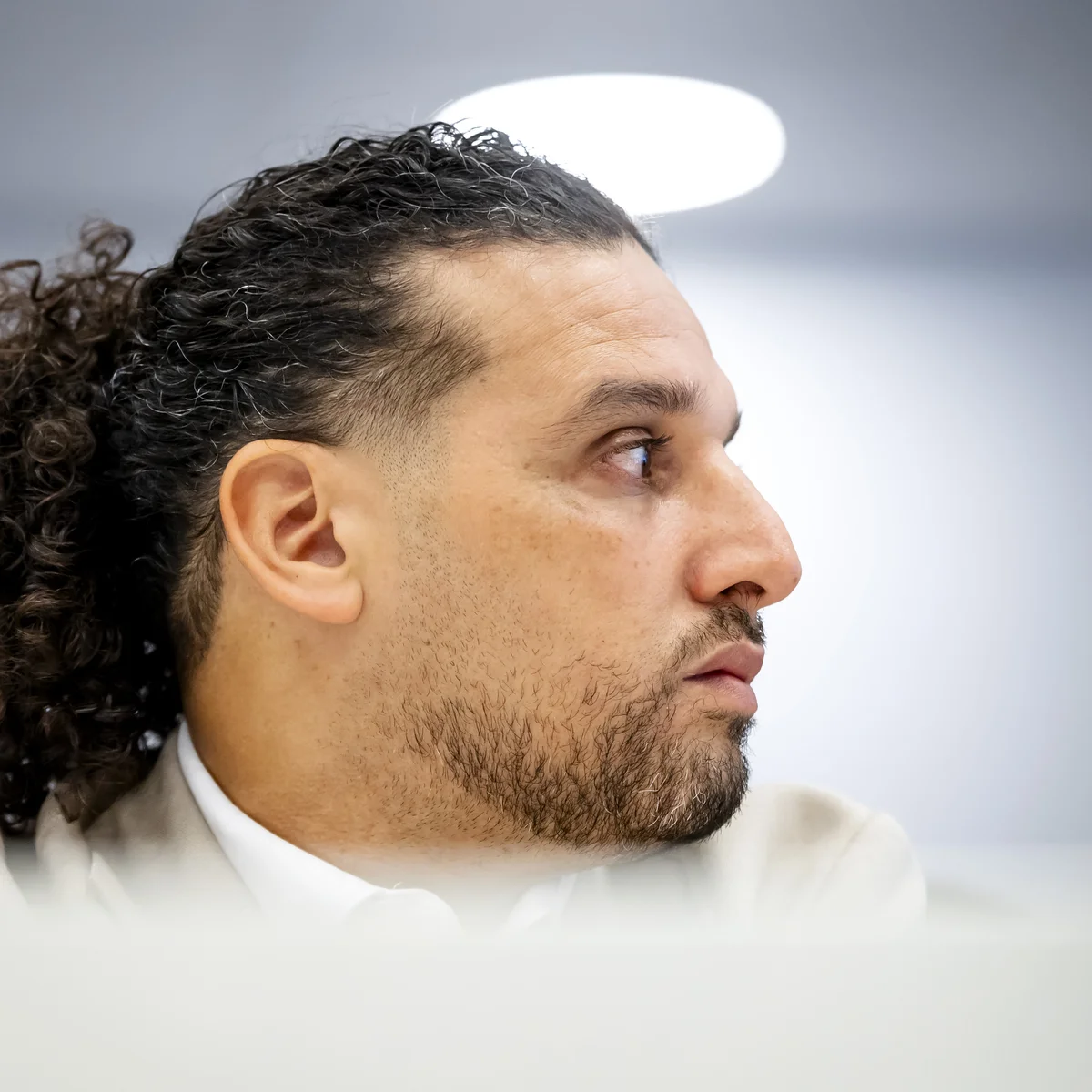 Profile view of a man with curly hair and a beard, looking intently to the side, in a softly lit room.