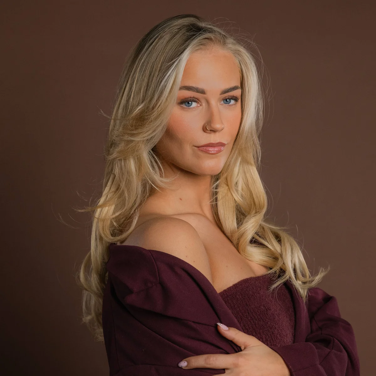 A woman Marijn Kuipers  with long blond hair and blue eyes poses confidently with a maroon off-shoulder sweater against a brown background.