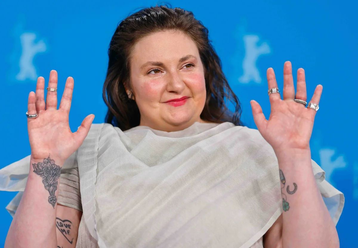 Person with tattoos raises hands, wearing a sheer white outfit, against a blue background. Smiling with rings on fingers.