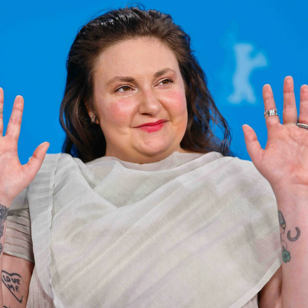 Person with tattoos raises hands, wearing a sheer white outfit, against a blue background. Smiling with rings on fingers.