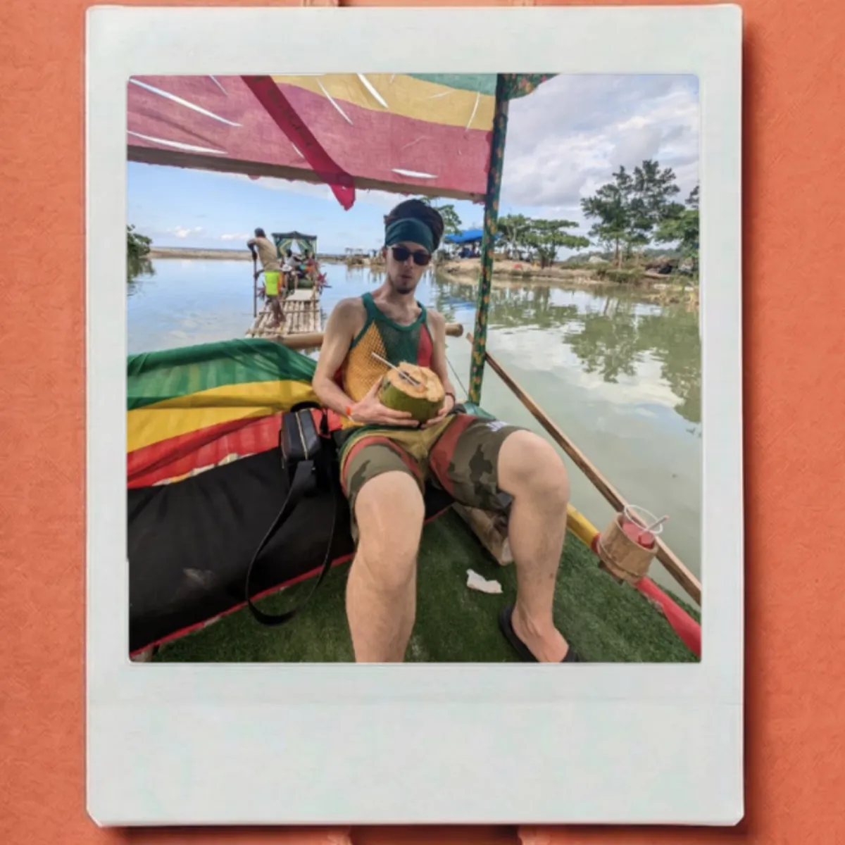 Person relaxing on a colorful floating raft, holding a coconut drink, beneath a bright canopy with trees and water in the background.