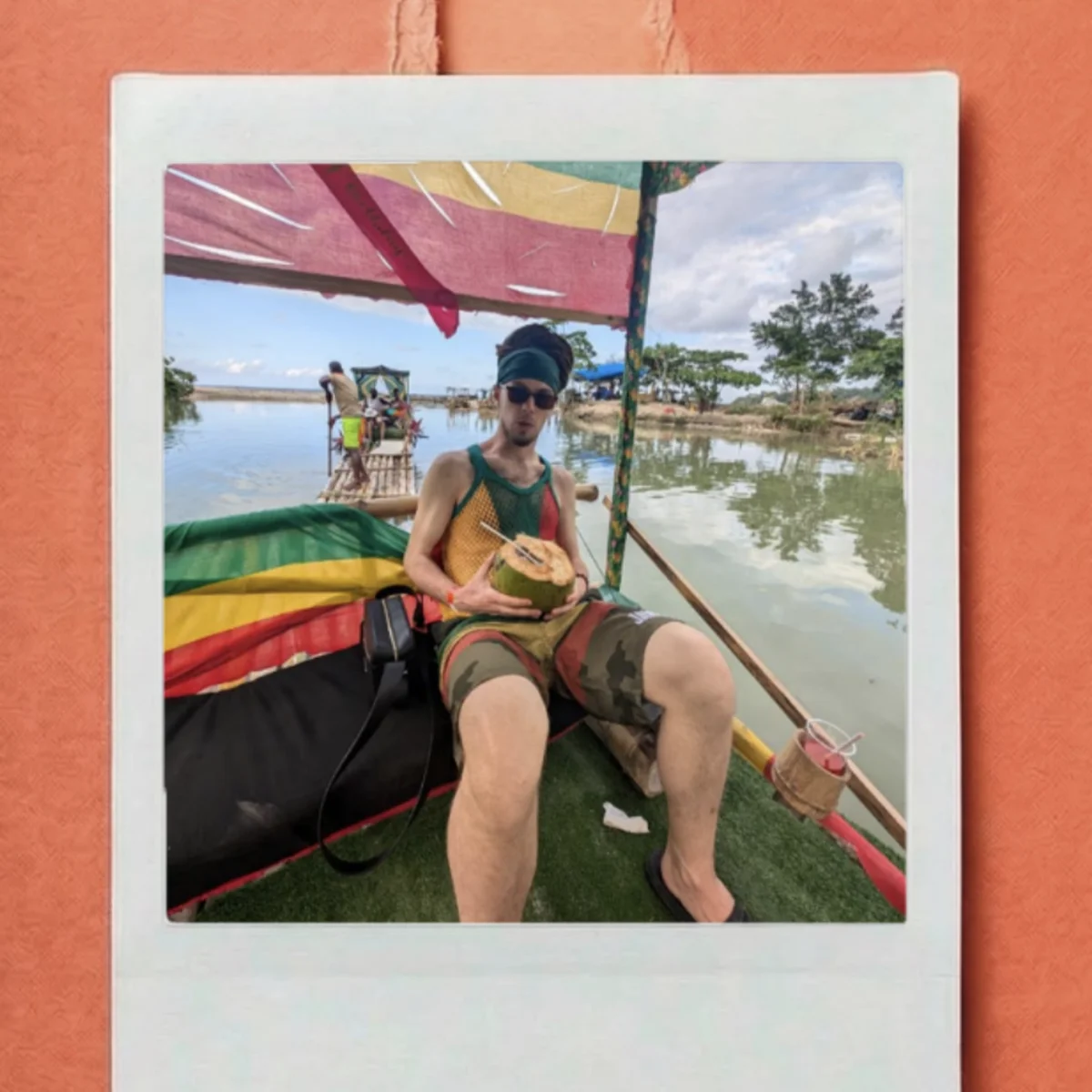 Person relaxing on a colorful floating raft, holding a coconut drink, beneath a bright canopy with trees and water in the background.