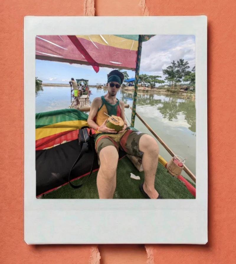 Person relaxing on a colorful floating raft, holding a coconut drink, beneath a bright canopy with trees and water in the background.