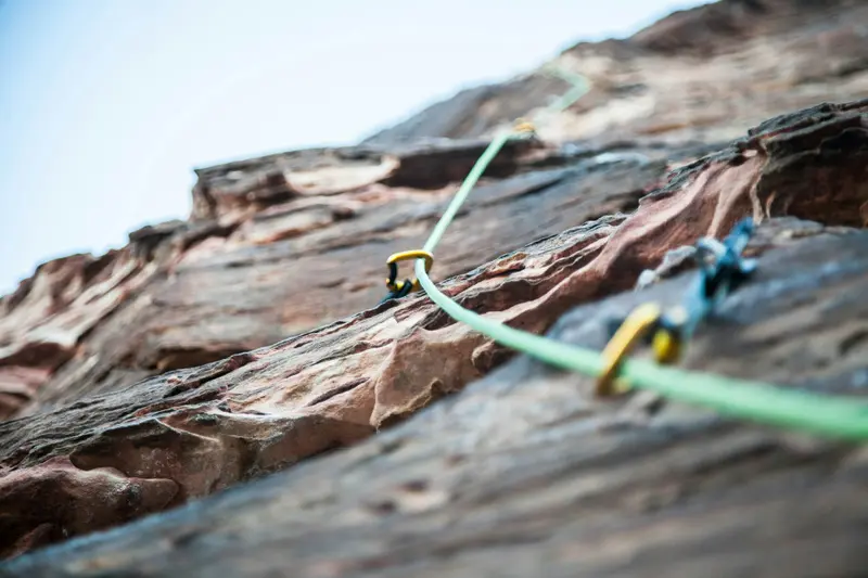 Close-up of a rock climbing wall with a green rope secured by carabiners leading to the top, emphasizing textured red-brown rock surface.