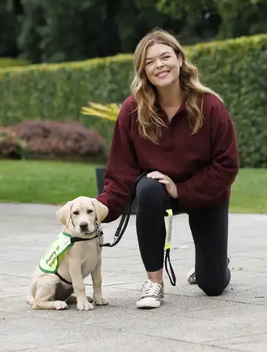 Doireann kneels beside a Labrador puppy wearing a "Puppy in Training" vest, smiling in a garden setting.