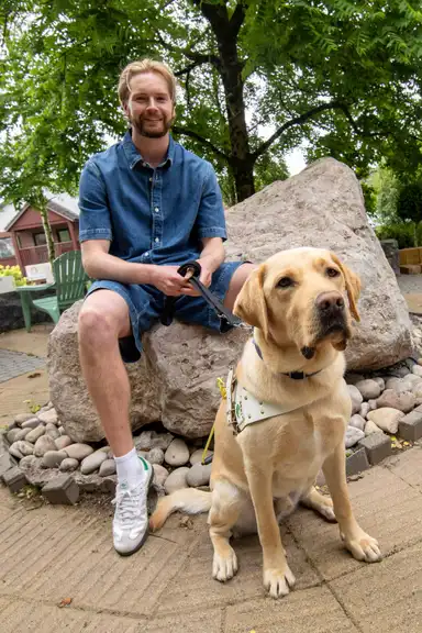 Caoimhín in a denim outfit sits on a rock, holding a leash attached to a guide dog. The dog sits obediently in a garden setting.