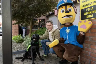 IGD Ambassador Shane Casey and a person in a Paw Patrol dog costume kneel beside a black working dog in a vest, outside the Training Centre with greenery and a "working dog" sign.