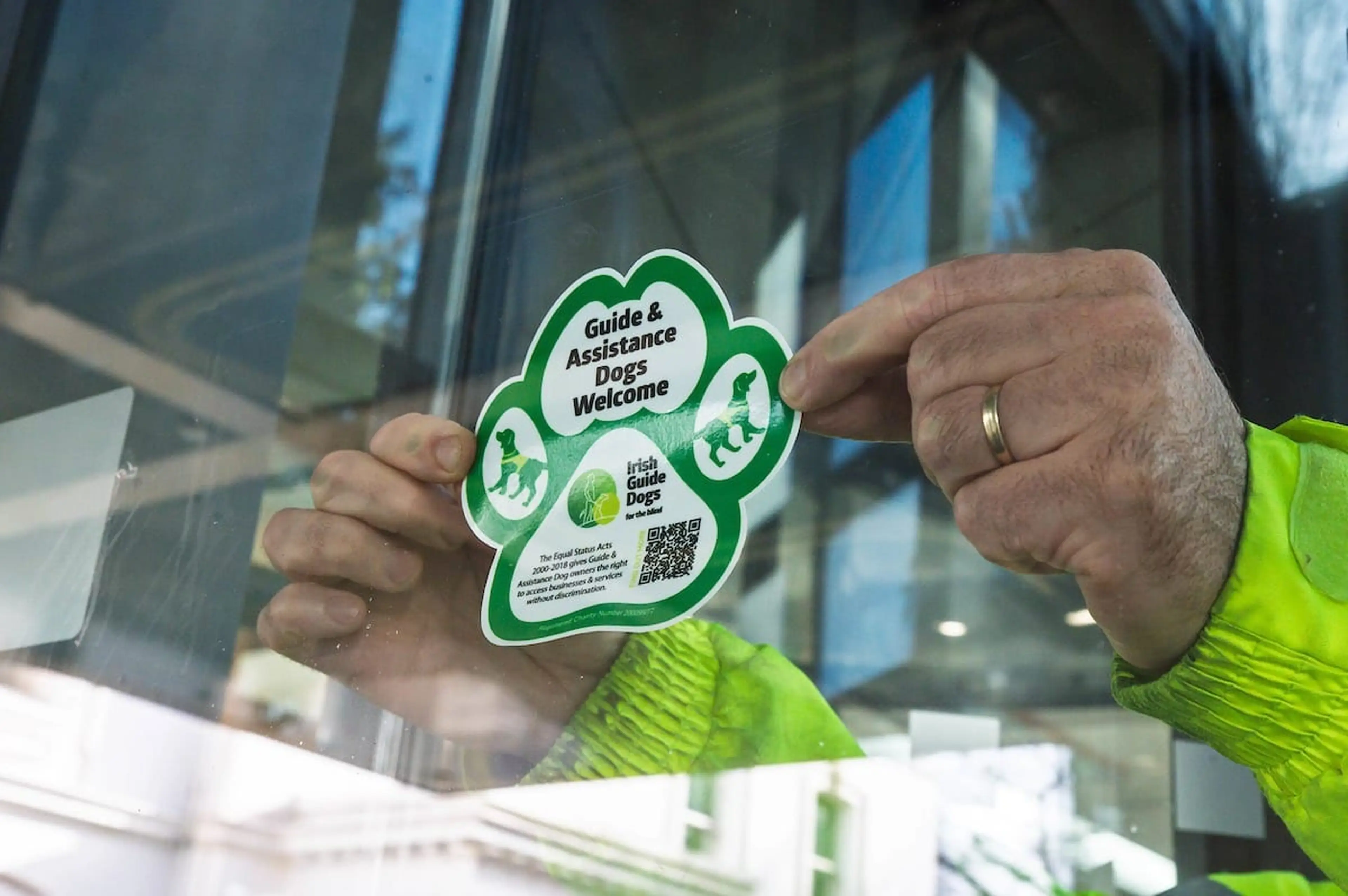Person placing a sticker on glass that says "Guide & Assistance Dogs Welcome." Sticker is shaped like a paw print with QR code and logos.