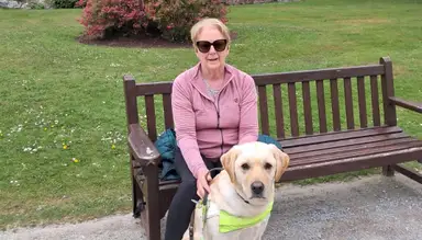 Geraldine in sunglasses sits on a park bench with a guide dog, surrounded by grass and flowers.