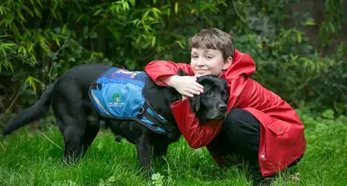 A boy in a red raincoat hugs a black Labrador wearing a blue service vest in a grassy garden setting.
