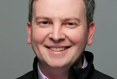A man with dark hair and a warm smile wearing a black jacket over a light purple shirt, photographed against a neutral gray background.