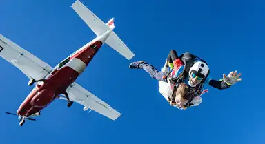 A skydiver in colorful gear freefalls alongside a red and white aircraft against a clear blue sky.