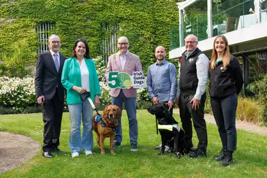 Board member Terence O'Keeffe, Assistance Dog owner Renaith Keane with Assistance Dog Kacie, Dublin Lord Mayor, Councillor Ray McAdam, Guide Dog owner Donnacha McCarthy with Guide Dog Ulysses, Tim O'Mahony (CEO of Irish Guide Dogs) and Puppy Raising Supervisor Frances Cooney all standing on grass as a group, with the Lord Mayor holding a "50 Irish Guide Dogs" sign, in front of a green ivy-covered building.