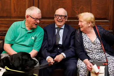 Tom Langan sitting in the middle with Guide Dog owner Tom O'Neill to his right and a woman to his left all sitting on a bench smiling and laughing, with Tom holding a black guide dog's leash.